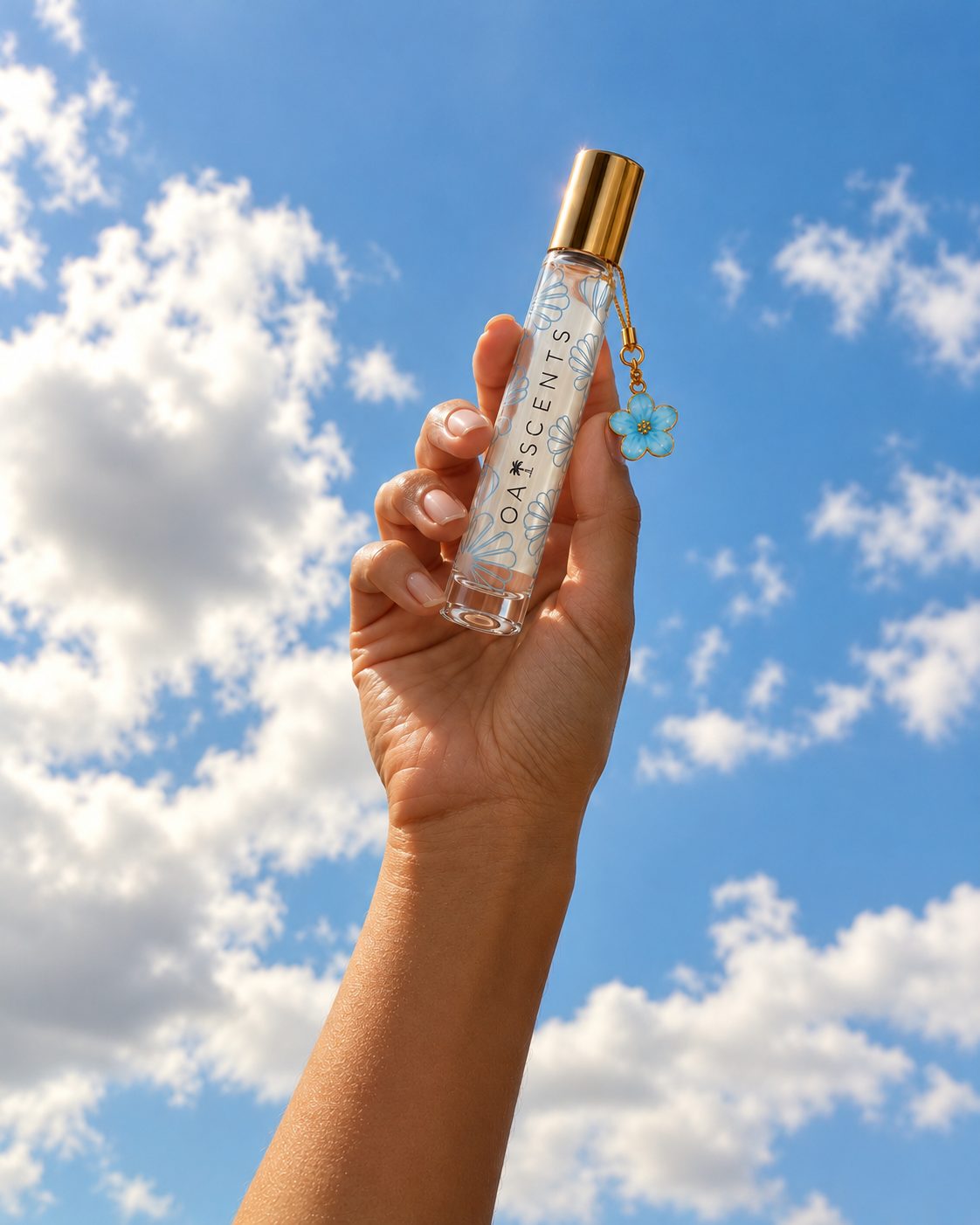 A hand lifting the Sky Bloom roll-on perfume oil against a bright blue sky, its gold cap and blue flower charm catching the light